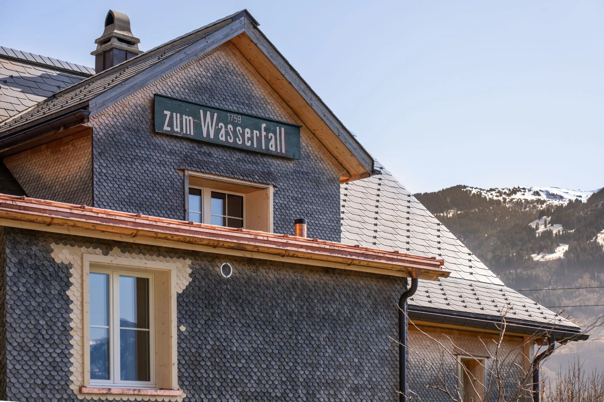 Schindelfassade mit dem Schild «zum Wasserfall» vor verschneiten Bergen