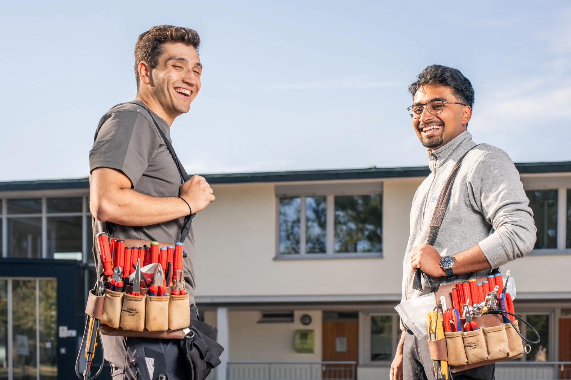 Zwei Elektriker von GalaTech mit Werkzeugtaschen lachen vor einem Gebäude in die Kamera